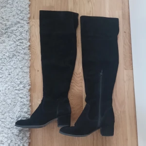 Svarta mockastövlar med klack - Snygga svarta stövlar i mocka med hög, över-knäet design och blockklack. De har rund tå och dragkedja på insidan för enkel påtagning. Perfekta för att lyfta din outfit under höst och vinter.