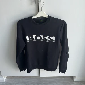 Svart BOSS sweatshirt med tryck - Svart sweatshirt från BOSS med stort vitt logotyptryck framtill. Klassisk rund halsringning och ribbade muddar vid ärmslut och nederkant. Skön passform och stilren design, perfekt för en clean och modern look. Köparen står för frakten!