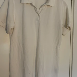 Klassisk vit pikétröja i bomull - En stilren vit pikétröja med korta ärmar och klassisk krage. Tröjan har knäppning framtill med två knappar och är gjord i mjuk bomull som känns skön mot huden. Perfekt för en clean och enkel look.