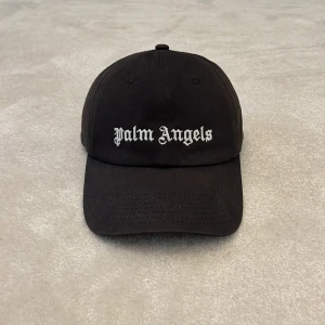 Svart keps från Palm Angels - Svart keps från Palm Angels med vit broderad logga både fram och bak. Klassisk böjd skärm och justerbart spänne bak. Tillverkad i 100% bomull med broderi i polyester. Perfekt accessoar för en streetwear-look. Det finns kvitto, tags och qr kod. Den är köpt på NK i Göteborg.