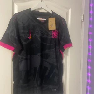 Chelsea x Nike svart fotbollströja - Chelsea x Nike Dri-FIT fotbollströja i svart med mörkgrått mönster, rosa detaljer på ärmslut och sidor samt gula inslag. Broderad rosa Chelsea-lejonlogga och Nike Swoosh framtill. Tillverkad i lätt och ventilerande material för maximal komfort.