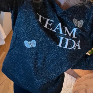 Glittrig svart tröja Ida Sjöstedt - Supersnygg svart glittrig tröja från Ida Sjöstedt med texten 'TEAM IDA' framtill. Tröjan har broderade fjärilar med pärldetaljer och en relaxed passform. Perfekt för dig som vill sticka ut lite extra med coola detaljer och glitter.