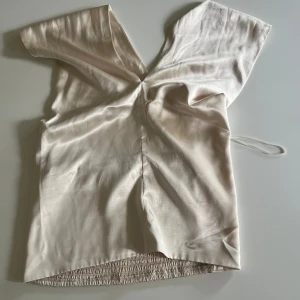 Champagnefärgad omlottopp från Bik Bok - Snygg champagnefärgad omlottopp från Bik Bok i glansigt satinliknande material. Toppen har korta ärmar, v-ringning och smockad rygg för extra passform. Banden på bild 3 är till för att knyta i nacken, så man får en öppen rygg🩷