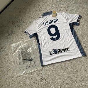 Inter Milan Thuram #9 Nike fotbollströja - Inter Milan bortatröja med Thuram och nummer 9 på ryggen. Vit med mörkblå detaljer, klubbmärke, sponsortryck och italienska mästarmärket på bröstet. Klassisk krage och korta ärmar. Tillverkad i lätt och ventilerande material, perfekt för match eller träning.