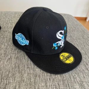 Svart New Era White Sox keps 59FIFTY - Snygg svart 59FIFTY keps från New Era med White Sox-logga framtill och broderade blå blommor. Kepsen har platt skärm, broderad World Series-patch på sidan och klassisk passform. Perfekt för dig som vill sticka ut med sportig stil.
