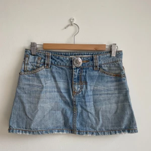 Ljusblå jeanskjol från ONLY JEANS  - Säljer en snygg ljusblå jeanskjol från ONLY med klassiska fickor fram och bak, synliga sömmar och rå kant nertill. Kjolen har bälteshällor och en stor metallknapp med logga framtill. Perfekt för dig som gillar en avslappnad och trendig stil.