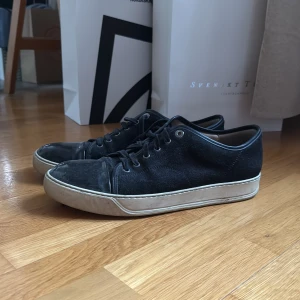 Svarta sneakers från Lanvin i mocka - Stilrena svarta sneakers från Lanvin med ovandel i mocka och detaljer i skinn. Skorna har rund tå, snörning och en vit platt sula som ger en clean kontrast. Perfekta för dig som gillar minimalistisk och lyxig streetstyle.