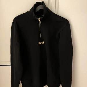 Svart tröja från Hugo Boss - Säljer en stilren svart tröja från Hugo Boss med dragkedja och hög krage. Tröjan har långärmade ärmar och en broderad logga på bröstet. Perfekt för en avslappnad men ändå elegant look.