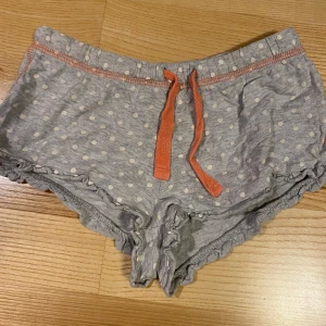 Pyjamas shorts strl 128 - Söta grå shorts med vita prickar och orange detaljer. Shortsen har en volangkant nedtill och ett orange snöre i midjan.