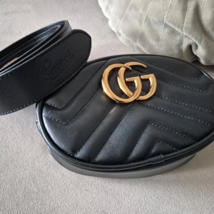 Svart quiltad midjeväska  - Snygg svart quiltad midjeväska från med emblem i guld på framsidan. Väskan har en dragkedja och är perfekt för att bära dina nödvändigheter med stil. Den justerbara remmen gör den bekväm att bära runt midjan.