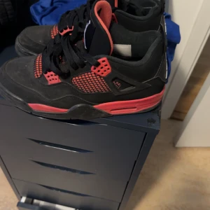 Jordan 4 red thunders - Snygga svarta och röda Nike Air Jordans med klassisk design. Skorna har snörning och en ikonisk logga på hälen. Perfekta för dig som vill ha en stilren och sportig look.