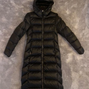 Svart dunjacka från Parajumpers - Säljer en lång svart dunjacka från Parajumpers med huva och dragkedja. Jackan är perfekt för kalla dagar och har en stilren design med quiltade detaljer. Endast använd ett fåtal gånger.