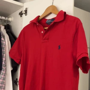 Röd pikétröja från Polo Ralph Lauren - Snygg röd pikétröja från Polo Ralph Lauren med klassisk krage och knappdetalj. Tröjan har en broderad logotyp i blått på bröstet och är tillverkad i bomull för en bekväm passform.