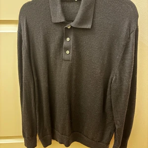 Svart pikétröja från arket - Snygg svart pikétröja med klassisk krage och tre knappar framtill. Perfekt för en stilren look. Tröjan har långa ärmar och är tillverkad i ett mjukt material som ger en bekväm passform.