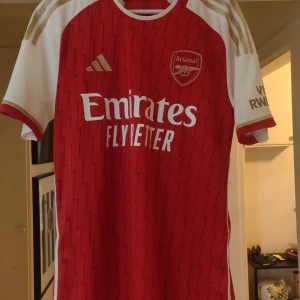 Röd Arsenal fotbollströja från Adidas - Snygg röd Arsenal fotbollströja från Adidas med vita detaljer och klubbens emblem på bröstet. Tröjan har korta ärmar och ett subtilt mönster över hela framsidan. Perfekt för alla Arsenal-fans! Aldrig använd vid fler bilder skriv gärna👍 finns ute på fler sidor