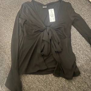 Oanvänd Svart knyt topp från Gina Tricot - Snygg svart långärmad topp från Gina Tricot med knytning framtill. Perfekt för en stilren look. Toppen har en v-ringad design och är tillverkad i ett mjukt material. Nypris 299