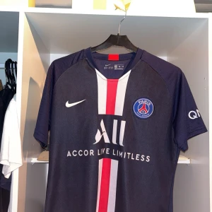 Blå PSG fotbollströja med Mbappé 7 - Snygg blå PSG-tröja från Nike med Mbappé och nummer 7 på ryggen. Tröjan har korta ärmar och ett rött och vitt vertikalt mönster på framsidan. Perfekt för fotbollsfans!