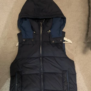 Mörkblå Puffer vest med luva - Säljer denna riktigt snygga mörkblåa vest med avtagbar luva. Vesten är i perfekt skick och det finns inga defekter fundera inte på att höra av dig😀
