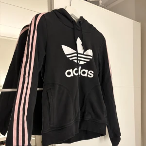 Svart hoodie från Adidas med rosa ränder - Säljer en svart hoodie från Adidas med den klassiska loggan i vitt på bröstet. Tröjan har långa ärmar med rosa ränder längs sidorna. Hör av er vid funderingar eller om ni vill ha bilder med hur plagget sitter på mig.