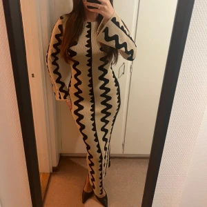 Beige och svart långklänning med zickzackmönster - Trendig långklänning i beige med svart zickzackmönster. Klänningen har långa ärmar och en figurnära passform som framhäver silhuetten. Jacquardstickad maxiklänning från nakd med utsvängda armar
