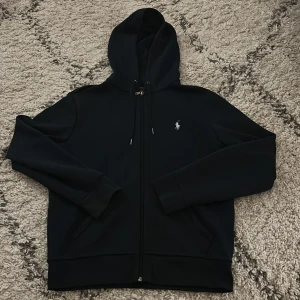 Svart hoodie från Ralph Lauren - Säljer en stilren svart hoodie från Ralph Lauren med dragkedja och huva. Den har en liten logotyp på bröstet och är perfekt för en avslappnad look. Passar bra till både jeans och träningsbyxor. Hör av dig vid intresse!