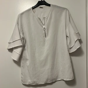 Vit blus från Lindex - Säljer en stilren vit blus från Lindex med dragkedja framtill och volangdetaljer på ärmarna. Perfekt för en chic och avslappnad look.