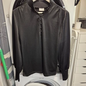 Blus - Blus I glansigt material med knappar upptill och muddar vid ärmsluten. Sparsamt använd 