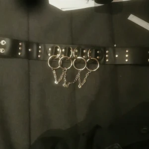 Svart halsband med kedjedetaljer - Coolt svart bälte med flera silverringar och kedjor som ger en edgy look. Bältet har nitar och är perfekt för att ge en extra touch till din outfit. Passar bra till både jeans och klänningar.