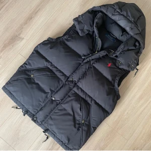 Svart dunväst från Polo Ralph Lauren - Snygg svart dunväst från Polo Ralph Lauren med en liten röd logga på bröstet. Västen har en avtagbar huva och stängs med dragkedja och knappar. 