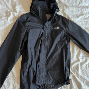 Svart regnjacka från The North Face - Säljer en svart regnjacka från The North Face. Jackan har en praktisk huva och dragkedja framtill. Perfekt för regniga dagar och håller dig torr och bekväm. En stilren och funktionell jacka för alla väder.
