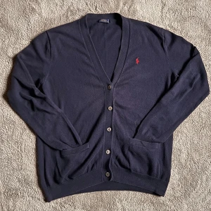 Ralph Lauren Cardigan - Säljer en stilren mörkblå kofta från Ralph Lauren med klassisk design. Koftan har en v-ringad öppning och knappar framtill. Den är dekorerad med det ikoniska röda Ralph Lauren-logotypen på bröstet. Perfekt för en elegant och avslappnad look. Storlek M och är true to size men funkar som en S också. 