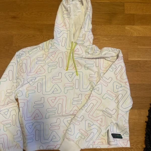 Vit hoodie från Fila med färgglatt mönster - Säljer en vit hoodie från Fila med ett färgglatt mönster i blått, rosa och grönt. Hoodien har en justerbar huva med gula snören och en bekväm passform. Perfekt för en avslappnad stil.