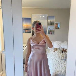 Ljusrosa sidenklänning - Elegant ljusrosa klänning i siden med smala axelband och en vacker omlott-design i ryggen. Klänningen har en mjuk och glansig yta som ger en lyxig känsla. Perfekt för speciella tillfällen.