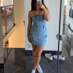 Jeansklänning i denim - Trendig axelbandslös jeansklänning i ljus denim. Perfekt för en avslappnad stil med en tight passform som framhäver figuren. Klänningen har en kort längd och är idealisk för varmare dagar.