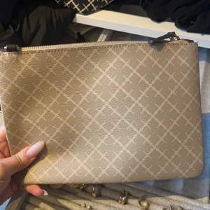 By Malene Birger väska beige  - Snygg beige kuvertväska med ett diskret rutigt mönster. Väskan har en dragkedja upptill och är perfekt för att bära med sig det nödvändigaste. Enkel och stilren design som passar till många tillfällen.