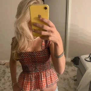 Rutig topp från Ganni - Säljer en snygg rutig topp från Ganni i rött och svart med smockdetaljer. Toppen har en croppad passform och breda axelband. Perfekt för en trendig look!