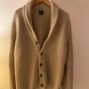 Beige kofta från Massimo Dutti - Säljer en stilren beige kofta från Massimo Dutti, tillverkad av italienskt garn. Koftan har en ribbad struktur och knappar framtill. Perfekt för en avslappnad men elegant look.