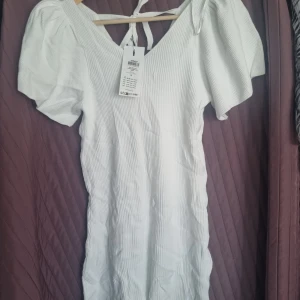 Vit ribbad klänning från Only - Säljer en stilren vit ribbad klänning från Only med korta puffärmar och v-ringning. Klänningen har en knytning i ryggen som ger en fin detalj. Perfekt för sommarens varma dagar!