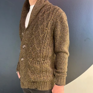 Brun stickad kofta med knappar - Säljer en snygg brun stickad kofta med knappar framtill. Koftan har ett flätmönster och ribbade muddar vid ärmslut och nederkant. Perfekt för kyliga dagar!