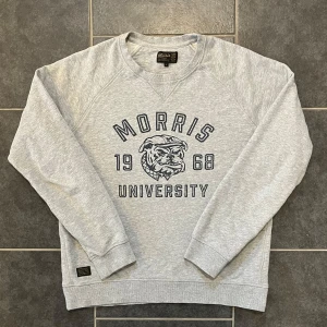 Grå sweatshirt från Morris - Säljer en grå sweatshirt från Morris med tryck 'Morris University 1968' och en bulldog på framsidan. Tröjan har långa ärmar och ribbade muddar vid ärmslut och nederkant. Perfekt för en avslappnad stil.