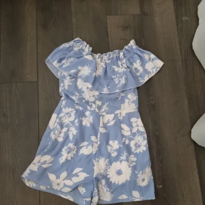 Blå blommig offshoulder klänning - Säljer en söt blå offshoulder klänning/dres med vita blommor. Klänningen har en volangdetalj upptill och är kort i modellen. Perfekt för sommarens varma dagar! 💙