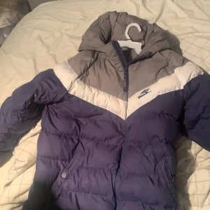 Blå och grå pufferjacka från Nike - Säljer en snygg blå och grå pufferjacka från Nike. Jackan har en dragkedja framtill och en huva för extra värme. Perfekt för kyliga dagar. Den har en cool design med kontrasterande färger och Nike-loggan på bröstet. Storleken passar dig som är mellan 158-170 cm lång.