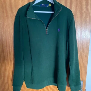 Grön tröja med dragkedja från Polo Ralph Lauren - Säljer en snygg grön tröja från Polo Ralph Lauren med dragkedja och lila broderad logga på bröstet. Perfekt för en stilren look. Tröjan har långa ärmar och en bekväm passform. Öppen för prisförslag