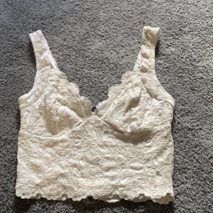 Vit spetsig linnetopp från Amisu XS - Superfin vit linnetopp från Amisu i storlek XS. Toppen är täckt av vacker spets med blommigt mönster och har breda axelband. Den är fodrad och har en lätt croppad passform med vågig kant nertill. Perfekt att styla med jeans eller kjol för en trendig look.