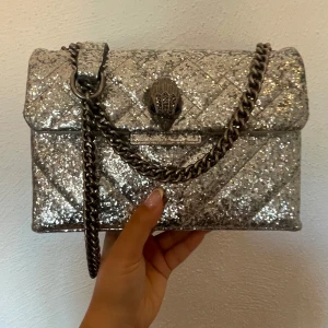 Glittrig silver väska från Kurt Geiger - Supersnygg väska från Kurt Geiger i glittrigt silver med quiltad yta och kedjerem. Väskan har ett coolt ormlås framtill och är perfekt för att sticka ut. Insidan är ljus och har flera fack för dina grejer. Kedjan är i metall 
