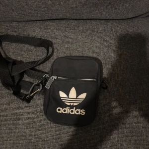 Svart axelväska från Adidas - Säljer en svart axelväska från Adidas med klassiska treklöver-loggan framtill och text på baksidan. Väskan har justerbar rem, dragkedja och ett huvudfack med innerficka. Perfekt storlek för mobil, plånbok och nycklar. Materialet är slitstark polyester.
