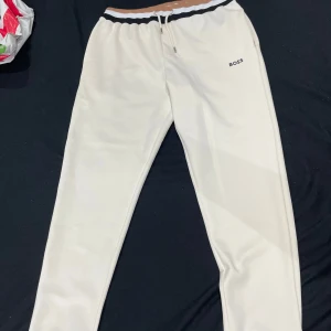 Vita mjukisbyxor från BOSS - Snygga vita mjukisbyxor från BOSS med svart och beige rand i midjan och diskret BOSS-logga på ena benet. Byxorna har dragsko i midjan och är tillverkade i ett mjukt, syntetiskt material som känns riktigt skönt mot huden och även. PRIS KAN DISKUTERAS