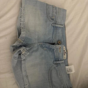 Ljusblå jeansshorts från Hollister - Snygga ljusblå jeansshorts från Hollister med fransiga kanter och klassisk femficksdesign. Shortsen har låg midja och är gjorda i mjukt denim med slitna detaljer, säljer då de är för stora