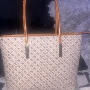 Beige shoppingväska med mönster - Snygg shoppingväska från Guess i beige med brunt logomönster över hela väskan. Väskan har långa bruna handtag, dragkedja upptill och flera innerfack. Perfekt storlek för att få plats med allt du behöver. Klassisk och stilren design med metall-detaljer.