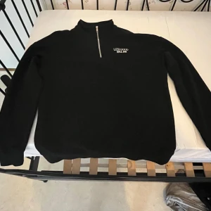 Svart half zip tröja Less Good - Svart långärmad tröja från Less Good med vit broderad text på bröstet och en snygg half zip-dragkedja framtill. Tröjan har en hög krage och är gjord i mjuk bomull, perfekt för chill dagar. Enkel och stilren design som funkar till jeans eller mjukis.